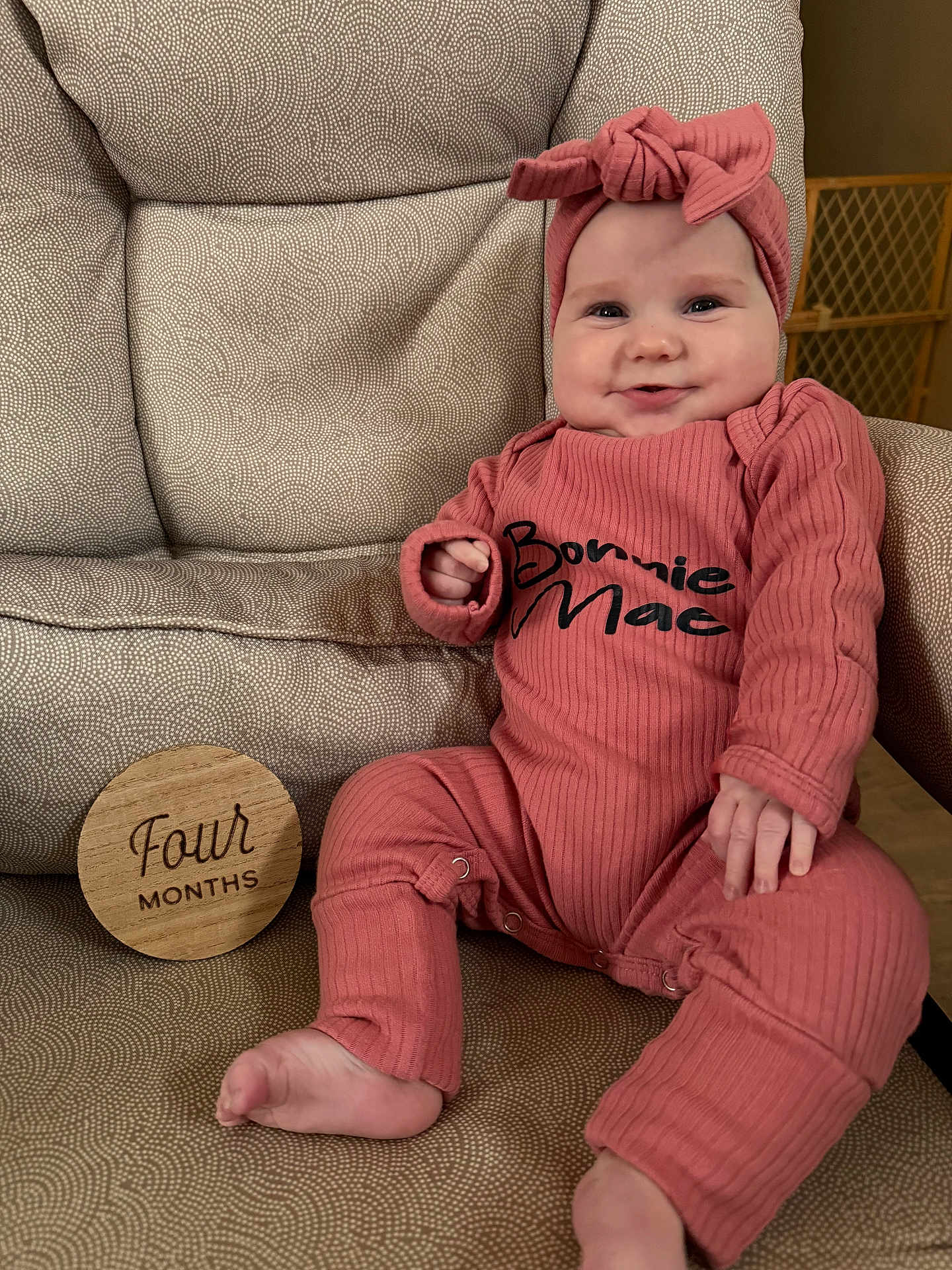 Bonnie joined the competition — help win amazing prizes! baby, infant, four_months, wooden_sign, headband, bow, pink_outfit, onesie, smile, chubby_cheeks, arm, leg, foot, chair, patterned_fabric, baby_month_marker, sitting, portrait, indoor, baby_gate