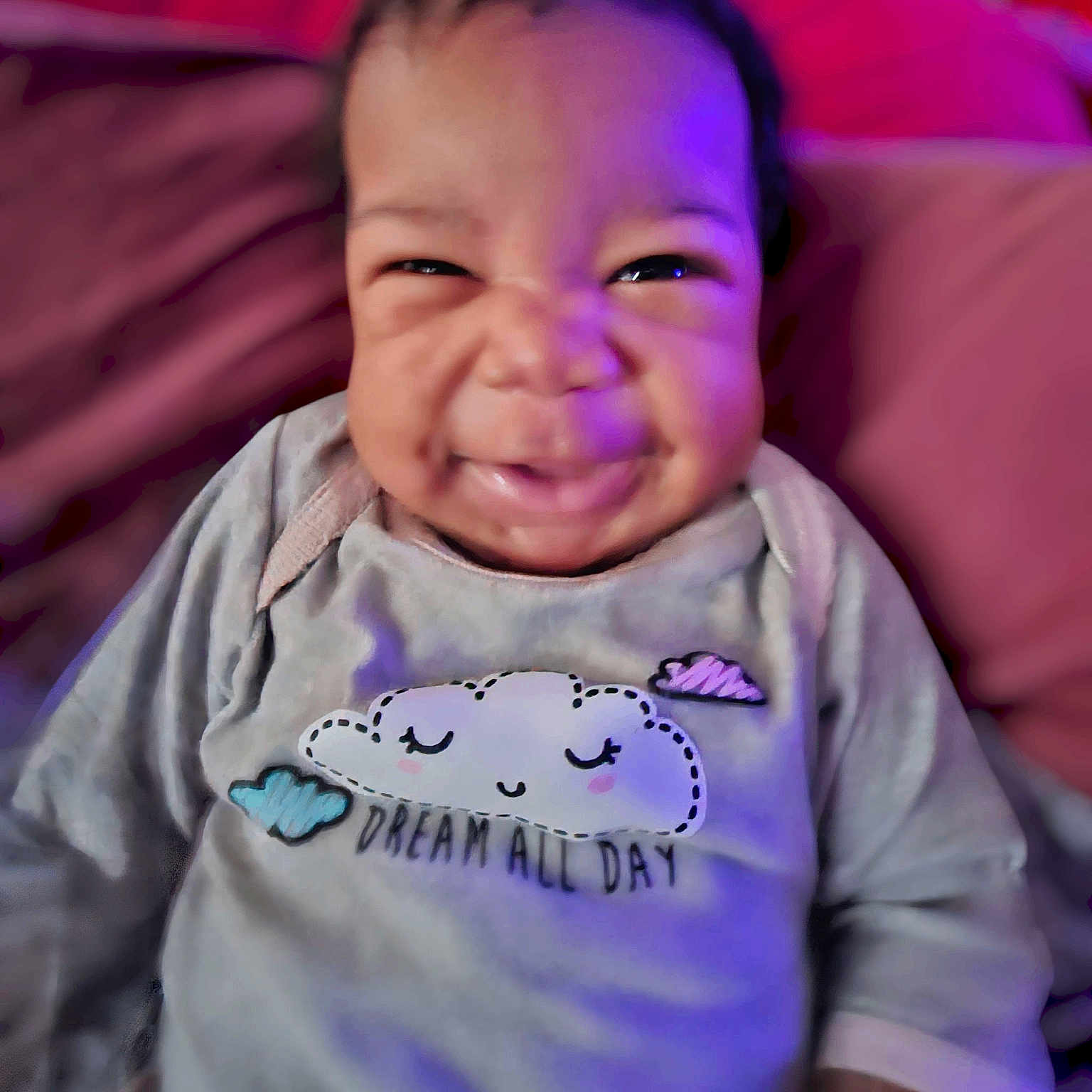 Amira is registered to the contest to win money with this photo: baby, candid, child, closeup, clothing, cozy, cute, dream, expression, face, happy, indoor, infant, long_sleeve, pillow, portrait, purple_light, sitting, smiling, soft_toys