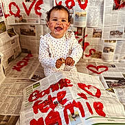Hélya participe au concours pour gagner de l'argent avec cette photo : toddler, child, newspaper, paint, red_paint, heart, smiling, sitting, white_clothing, curly_hair, face, hands, indoor, celebration, messy, art_project, writing, background, floor, happy