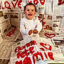 toddler, child, newspaper, paint, red_paint, heart, smiling, sitting, white_clothing, curly_hair, face, hands, indoor, celebration, messy, art_project, writing, background, floor, happy
