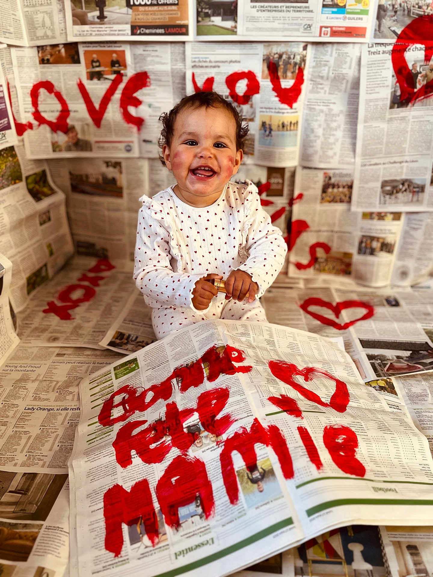 Hélya participe au concours pour gagner de l'argent avec cette photo : toddler, child, newspaper, paint, red_paint, heart, smiling, sitting, white_clothing, curly_hair, face, hands, indoor, celebration, messy, art_project, writing, background, floor, happy