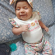 Abigail is registered to the contest to win money with this photo: baby, smiling, floral_outfit, headband, pacifier, bedspread, patterned, child, cute, happy, infant, lying_down, clothing, white_top, colorful, indoors, person, closeup, resting, adorable