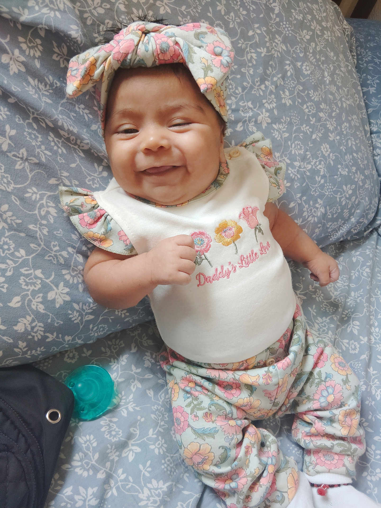 Abigail is registered to the contest to win money with this photo: baby, smiling, floral_outfit, headband, pacifier, bedspread, patterned, child, cute, happy, infant, lying_down, clothing, white_top, colorful, indoors, person, closeup, resting, adorable