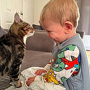 Ralph joined the competition — help win amazing prizes! baby, cat, tabby_cat, child, couch, indoor, curious, smiling, face, hands, clothing, blanket, home, furniture, interaction, cute, playful, portrait, soft_light, cozy