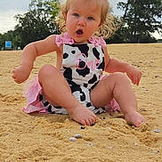 Aubrey joined the competition — help win amazing prizes! toddler, child, sand, outdoor, beach, cow_print, pink, dress, barefoot, curly_hair, expression, nature, trees, cloudy_sky, cute, sitting, playful, young_child, person, summer