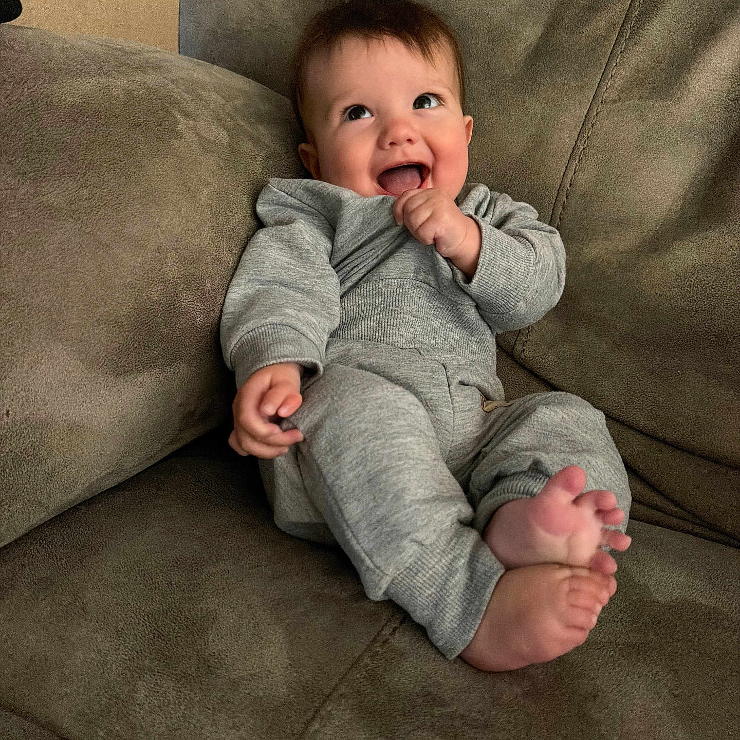 Beau is registered to the contest to win money with this photo: baby, child, smiling, happy, couch, sofa, indoor, clothing, grey_outfit, feet, hand, face, head, person, infant, seated, cute, expression, joy, cozy