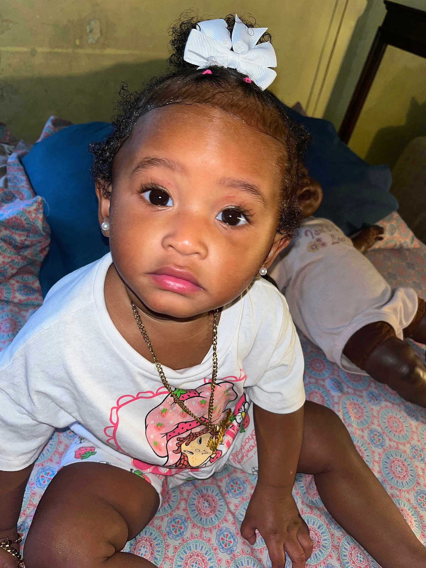 Royalty is registered to the contest to win money with this photo: toddler, child, baby, white_bow, strawberry_shortcake, gold_chain, earrings, curly_hair, patterned_blanket, doll, indoor, cute, portrait, sitting, clothing, person, face, eyes, hand, toy