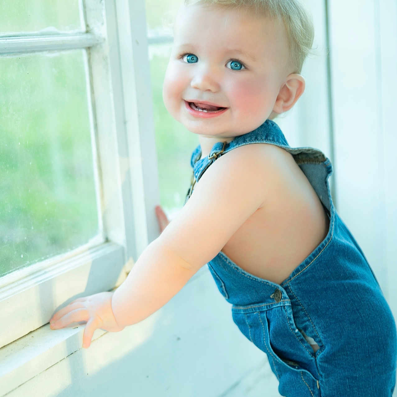 Stetson is registered to the contest to win money with this photo: baby, blond_hair, blue_eyes, casual_clothing, cheeks, child, cute, denim_overalls, face, hands, happy, home, indoor, natural_light, portrait, skin, smiling, standing, toddler, window