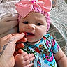 baby, bed, blanket, child, colorful_clothing, curious, cute, expression, feeding, food, hand, headband, indoor, infant, person, pink_bow, portrait, skeleton_print, tattoo, tropical_print