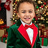 child, smiling, holiday_clothing, green_jacket, red_bow_tie, christmas_tree, christmas_decorations, wrapped_presents, festive, holiday, indoors, portrait, happy, celebration, seasonal, young_child, curly_hair, formal_wear, background_bokeh, festive_spirit