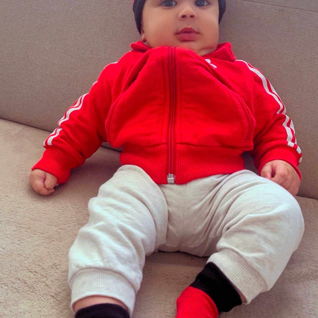 Alseny Camara participe au concours pour gagner de l'argent avec cette photo : baby, child, infant, red_jacket, hat, socks, couch, indoor, casual_clothing, cute, person, seated, relaxed, home, portrait, looking_at_camera, comfortable, young_child, fashion, sporty