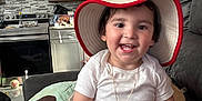 Erick joined the competition — help win amazing prizes! toddler, child, hat, smile, white_clothing, necklace, plush_toy, couch, living_room, kitchen, cabinet, appliance, floor, happy, sitting, indoor, person, furniture, home, casual