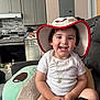 toddler, child, hat, smile, white_clothing, necklace, plush_toy, couch, living_room, kitchen, cabinet, appliance, floor, happy, sitting, indoor, person, furniture, home, casual