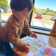 Erick joined the competition — help win amazing prizes! toddler, child, sweater, brown, activity_sheet, table, window, parking_lot, car, blue_sky, indoor, daylight, hair, face, hands, furniture, person, casual_clothing, reading, playing