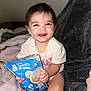 baby, child, smile, snack, food, package, blanket, sitting, indoors, cute, happy, toddler, clothing, person, face, hand, furniture, soft, comfort, playful