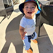 Maximiliano joined the competition — help win amazing prizes! toddler, child, smiling, hat, bucket_hat, casual_clothing, outdoor, sunlight, shadow, wicker_chair, patio, shoes, happy, reaching_hand, concrete_floor, daylight, person, cute, young_child, playful