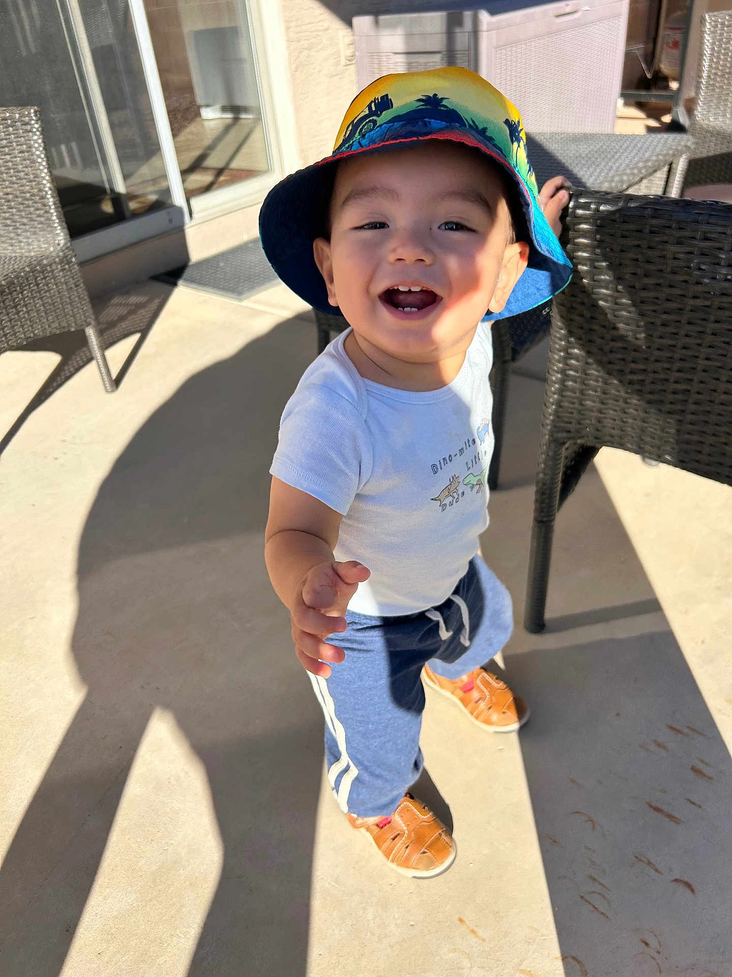 Maximiliano joined the competition — help win amazing prizes! toddler, child, smiling, hat, bucket_hat, casual_clothing, outdoor, sunlight, shadow, wicker_chair, patio, shoes, happy, reaching_hand, concrete_floor, daylight, person, cute, young_child, playful
