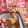 baby, balloons, birthday, cake, celebration, child, crown, decorations, first_birthday, flowers, gold, indoors, neon_sign, pacifier, party, pink, playpen, smiling, table, toys