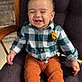 baby, child, smiling, laughing, plaid_shirt, flower, yellow_flower, pants, rust_color, chair, cushion, indoor, wooden_armrest, happy, cute, portrait, seated, infant, person, clothing