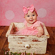 Everlee is registered to the contest to win money with this photo: baby, bow, child, cute, decor, face, floor, flower_garland, happy, headband, indoor, infant, pink_background, pink_dress, portrait, sitting, smile, toddler, wood, wooden_drawer