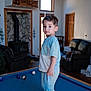Idrees is registered to the contest to win money with this photo: toddler, child, pool_table, living_room, blue_socks, smiley_face, wooden_beams, wood_stove, leather_couch, window, door, indoor, playing, casual_clothing, carpet, furniture, home_interior, curly_hair, expression, standing