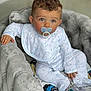 baby, child, pacifier, pajamas, armchair, curly_hair, blue_eyes, plush_furniture, indoor, comfort, infant, cute, soft_toys, clothing, sitting, portrait, young_child, baby_bib, relaxed, home