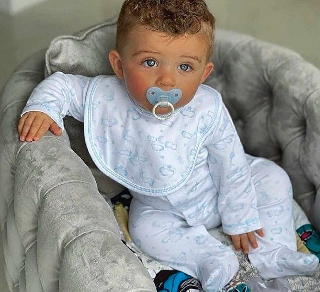 Kayden participe au concours pour gagner de l'argent avec cette photo : baby, child, pacifier, pajamas, armchair, curly_hair, blue_eyes, plush_furniture, indoor, comfort, infant, cute, soft_toys, clothing, sitting, portrait, young_child, baby_bib, relaxed, home
