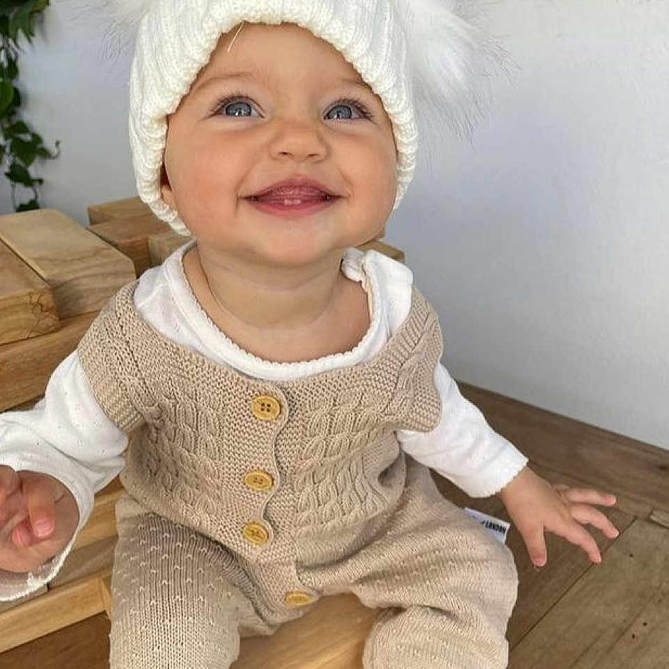 Maïlyna participe au concours pour gagner de l'argent avec cette photo : baby, blue_eyes, button, child, clothing, cozy, cream, cute, face, hand, happy, hat, indoor, infant, knitwear, portrait, sitting, smiling, white, wood