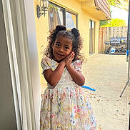 Jazlynn is registered to the contest to win money with this photo: child, girl, curly_hair, dress, butterfly_pattern, posing, smiling, indoors, glass_door, backyard, grill, concrete_floor, sunlight, window, hair_puffs, hands, happy, young, person, portrait