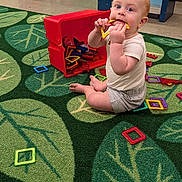 Theo joined the competition — help win amazing prizes! baby, child, infant, sitting, smiling, face, eyes, short_hair, indoor, classroom, daycare, playroom, carpet, toy, magnetic_tiles, red_bin, shelf, play_table, colorful, portrait