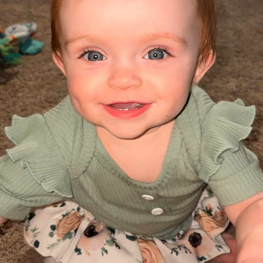 Bailee is registered to the contest to win money with this photo: baby, blue_eyes, carpet, child, clothing, cute, floor, floral_skirt, green_top, happy, home, indoor, person, playroom, portrait, reaching, ruffles, smiling, toddler, young_child
