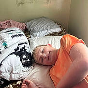 Peaches is registered to the contest to win money with this photo: boy, cat, bed, blanket, pillow, indoor, resting, relaxed, child, pet, feline, orange_shirt, black_and_white_cat, face, carpet, cozy, lying_down, blanket_pattern, wall, casual