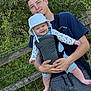 Elsie is registered to the contest to win money with this photo: baby, bucket_hat, baby_carrier, young_man, outdoor, greenery, wooden_fence, smiling, happy, person, nature, casual_clothing, holding, portrait, daylight, trees, grass, face, parenting, bonding