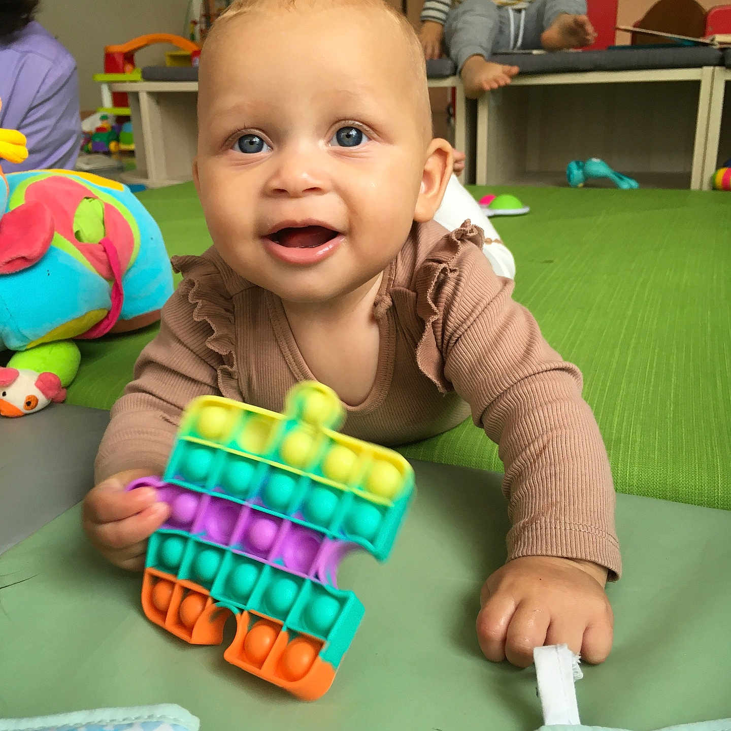 Talyssa a rejoint le concours — aidez-le/la à gagner de superbes lots ! baby, background, blue_eyes, child, clothing, colorful, face, floor, furniture, green_mat, hand, happy, indoor, person, play, playroom, pop_it, smiling, toddler, toy