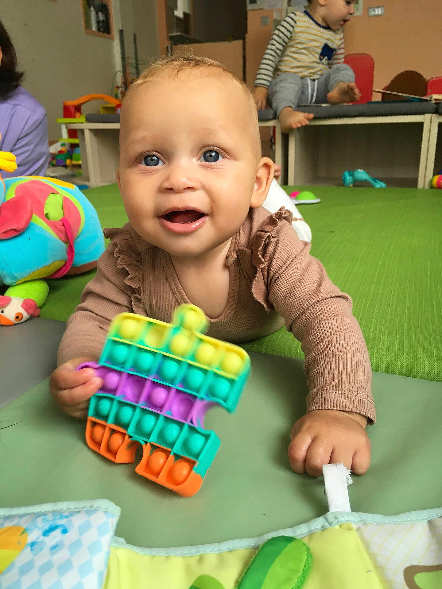 Talyssa a rejoint le concours — aidez-le/la à gagner de superbes lots ! baby, child, toddler, playroom, toy, pop_it, colorful, blue_eyes, green_mat, clothing, person, indoor, happy, smiling, hand, face, play, floor, background, furniture