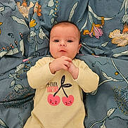 Eliana participe au concours pour gagner de l'argent avec cette photo : baby, blanket, blue_background, cherry_print, child, clothing, comfortable, cute, expression, face, floral_pattern, hands, indoors, infant, lying_down, onesie, person, portrait, soft, young