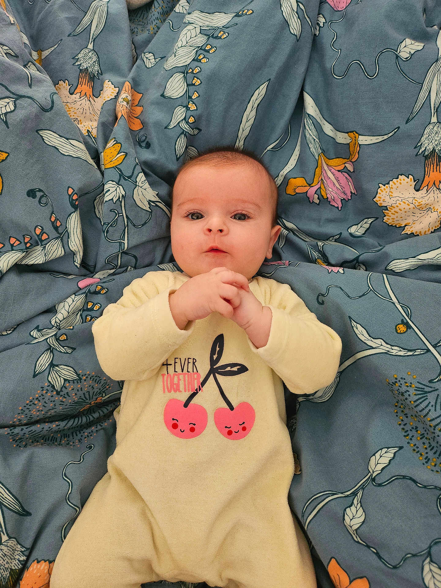 Eliana participe au concours pour gagner de l'argent avec cette photo : baby, blanket, blue_background, cherry_print, child, clothing, comfortable, cute, expression, face, floral_pattern, hands, indoors, infant, lying_down, onesie, person, portrait, soft, young