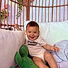 Lucca is registered to the contest to win money with this photo: baby, child, smiling, onesie, couch, plush_toy, green_alligator, purple_cushion, wicker_chair, indoor, happy, infant, cute, soft_toys, furniture, person, relaxing, home, casual, seated