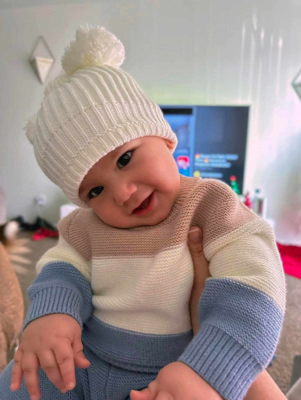 Matteo joined the competition — help win amazing prizes! baby, child, knitted_hat, pom_pom, sweater, striped_clothing, smile, face, hand, indoor, living_room, blurred_background, television, carpet, cozy, cute, person, infant, warm_clothing, portrait