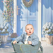 Brighton a rejoint le concours — aidez-le/la à gagner de superbes lots ! baby, child, wheelbarrow, blue_door, wreath, flowers, plants, striped_jacket, wooden, garden, cute, portrait, expression, outdoor, decor, potted_plants, greenery, door_handle, bright, seated