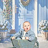 baby, child, wheelbarrow, blue_door, wreath, flowers, plants, striped_jacket, wooden, garden, cute, portrait, expression, outdoor, decor, potted_plants, greenery, door_handle, bright, seated