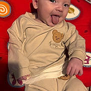 George joined the competition — help win amazing prizes! baby, child, infant, tongue_out, smiling, pajamas, teddy_bear, red_cushion, patterned_cushion, cozy, cute, indoors, portrait, playful, hand, face, head, person, soft_lighting, relaxed