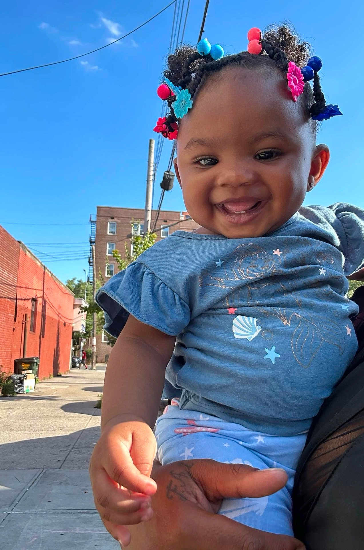 Ta'Sani is registered to the contest to win money with this photo: child, toddler, smiling, outdoor, blue_sky, hair_beads, urban, street, holding_hand, clothing, person, happy, daylight, building, sidewalk, tattoo, cute, portrait, baby, casual