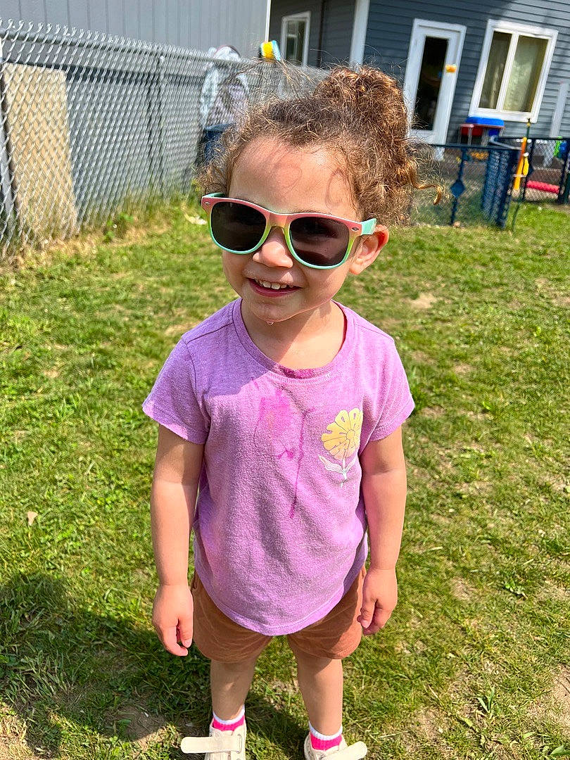 Kiara is registered to the contest to win money with this photo: baby_toddler_clothing, cool, eyewear, fence, glasses, goggles, grass, happy, joy, person, pink, plant, purple, recreation, shorts, sleeve, smile, summer, sunglasses, toddler
