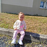Mya a rejoint le concours — aidez-le/la à gagner de superbes lots ! toddler, child, sitting, outdoor, grass, curb, sweatshirt, pink_pants, white_shoes, cartoon_character, building, window, sunlight, shadow, concrete, gravel, smile, person, young_child, casual_clothing