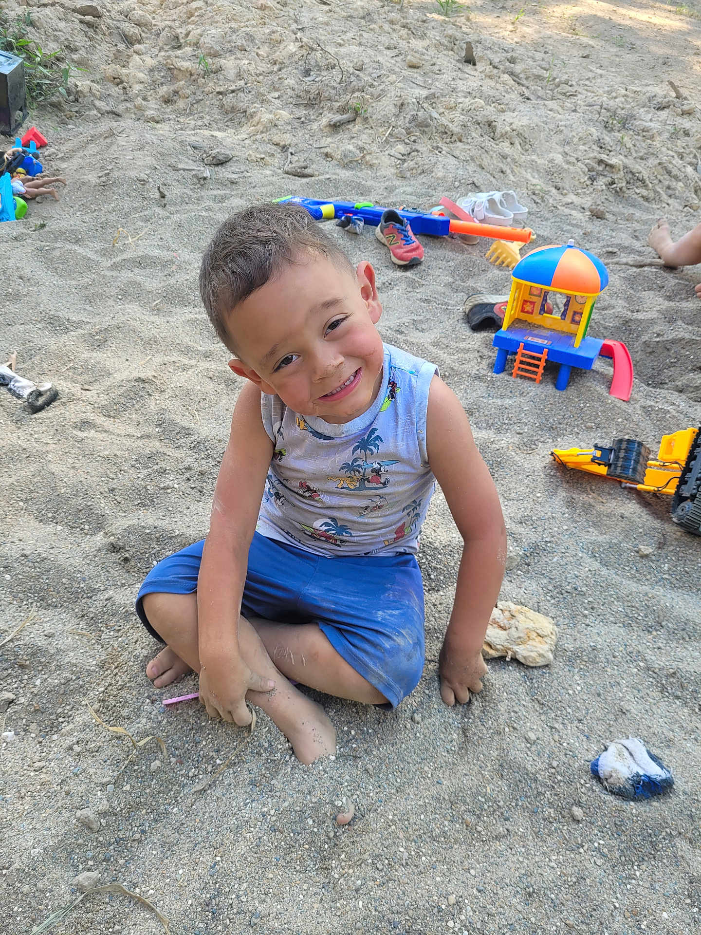 Luka is registered to the contest to win money with this photo: child, boy, sand, outdoor, play, toys, smile, happy, barefoot, shorts, tank_top, dirt, playground, summer, fun, casual_clothing, sitting, messy, daylight, nature