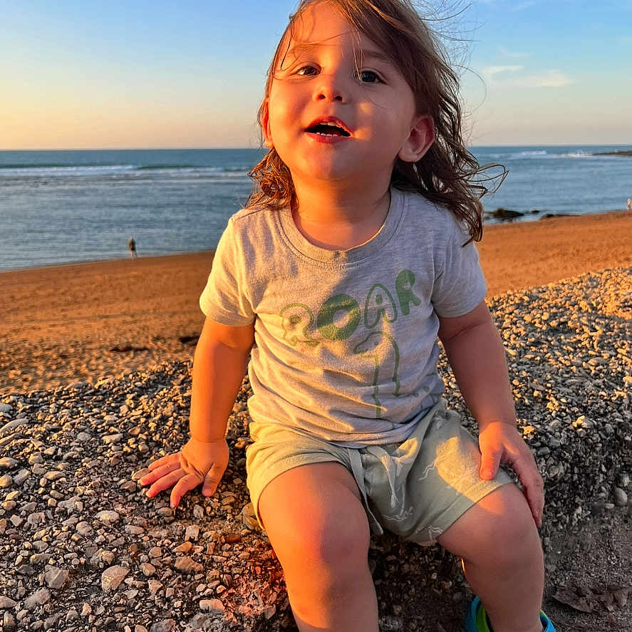 Hollyster participe au concours pour gagner de l'argent avec cette photo : toddler, child, beach, ocean, sunset, rocks, tshirt, shorts, shoes, smiling, outdoor, sunlight, portrait, nature, sky, water, sand, happy, casual_clothing, wind