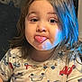 child, toddler, tongue_out, long_hair, indoor, shirt, car_pattern, food_stain, blue_light, playful, cute, face, expression, person, young_child, smile, seated, casual_clothing, portrait, funny