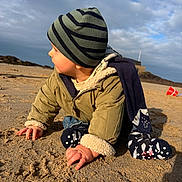 Malo participe au concours pour gagner de l'argent avec cette photo : toddler, child, beach, sand, hat, jacket, crawling, outdoor, cloudy_sky, sunlight, warm_clothing, bucket, toy, nature, play, portrait, side_profile, person, winter_clothes, daytime