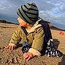 toddler, child, beach, sand, hat, jacket, crawling, outdoor, cloudy_sky, sunlight, warm_clothing, bucket, toy, nature, play, portrait, side_profile, person, winter_clothes, daytime